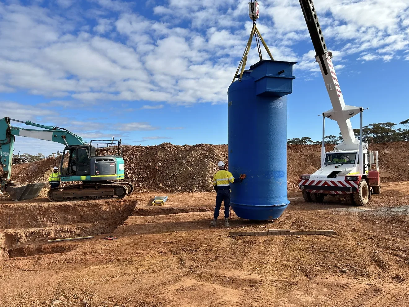 A blue tank being lifted into an excavation by crane on a red-dirt site